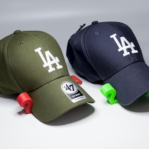 May include: Two baseball caps, one olive green and one navy blue, each with a white "LA" logo. The caps are displayed on red and green supports, with a "47 MVP" adjustable tag visible on one.