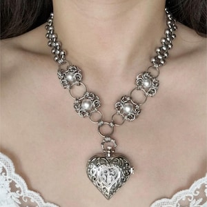 May include: Silver-toned necklace featuring a heart-shaped pendant with intricate filigree detailing. The necklace has a chain of small, round beads and decorative floral elements with pearl accents. The pendant is suspended from a series of connecting rings.