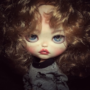 May include: A close-up of a doll with light skin, blue eyes, and curly brown hair. The doll has rosy cheeks and lips. It is wearing a gray long-sleeved shirt with a black collar and a pattern of black eyeglasses.
