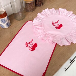 May include: A pink kitchen set featuring a rectangular mat with a red border and a round, ruffled placemat. Both items have a red cherry embroidery. The set is displayed on a wooden surface.