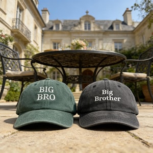 Puede incluir: Se muestran dos gorras de béisbol, una verde oscuro y otra gris antracita. La gorra verde dice "BIG BRO" en blanco, mientras que la gorra gris dice "Big Brother". Las gorras están sobre una superficie de piedra, con una mesa y sillas al fondo.