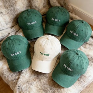 Puede incluir: Seis gorras de béisbol. Cinco son de color verde oscuro con "THE GIRLS" bordado en blanco. Una es de color blanco roto con "THE BRIDE" bordado en verde. Las gorras están sobre una superficie de piel sintética.