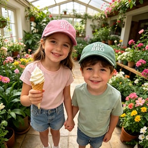 Pode incluir: Duas crianças usando bonés de beisebol combinando com os textos "BIG SIS" e "BIG BRO". A menina segura um sorvete. Eles sorriem e dão as mãos em uma estufa cheia de flores.