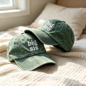 Puede incluir: Dos gorras de béisbol de color verde salvia con texto bordado en blanco. Una gorra presenta el texto "big bro" y la otra el texto "big sis". Ambas gorras tienen un aspecto desgastado, una visera curva y ojales cosidos en la corona.