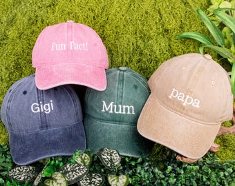 May include: Four baseball caps in pink, navy, green, and tan. Each cap has white embroidered text: "Fun Fact!", "Gigi", "Mum", and "papa". The caps are arranged on a bed of green foliage.