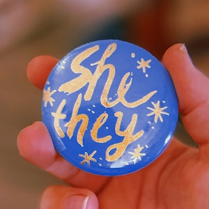 Könnte beinhalten: Ein runder blauer Knopf mit goldener Beschriftung, auf dem "she*they*" mit goldenen Sternen um die Beschriftung steht.