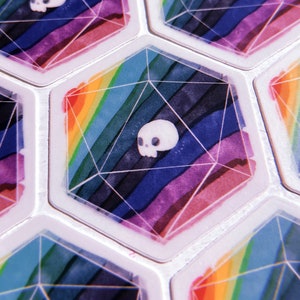 Puede incluir: Primer plano de fichas de juego hexagonales con un dise&ntilde;o de arco&iacute;ris y una calavera blanca en el centro de cada ficha.