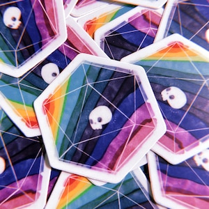 Puede incluir: Primer plano de una pila de pegatinas de arco&iacute;ris de colores con un dise&ntilde;o de calavera dentro de una forma geom&eacute;trica. Las pegatinas est&aacute;n impresas en papel blanco y tienen un acabado brillante.