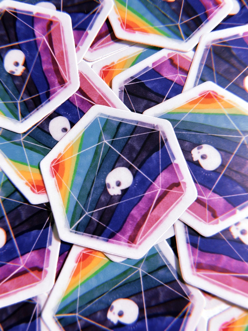Puede incluir: Primer plano de una pila de pegatinas de arco&iacute;ris de colores con un dise&ntilde;o de calavera dentro de una forma geom&eacute;trica. Las pegatinas est&aacute;n impresas en papel blanco y tienen un acabado brillante.