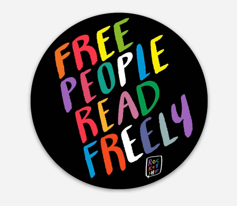 May include: A black and white round sticker with the words "FREE PEOPLE READ FREELY" written in a rainbow of colors. The words are written in a diagonal pattern and the text "ROCKET SHIP" is in a small speech bubble at the bottom.