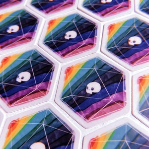 Puede incluir: Primer plano de fichas de juego hexagonales con un dise&ntilde;o de arco&iacute;ris y un gr&aacute;fico de calavera. Las fichas son blancas con un acabado brillante.