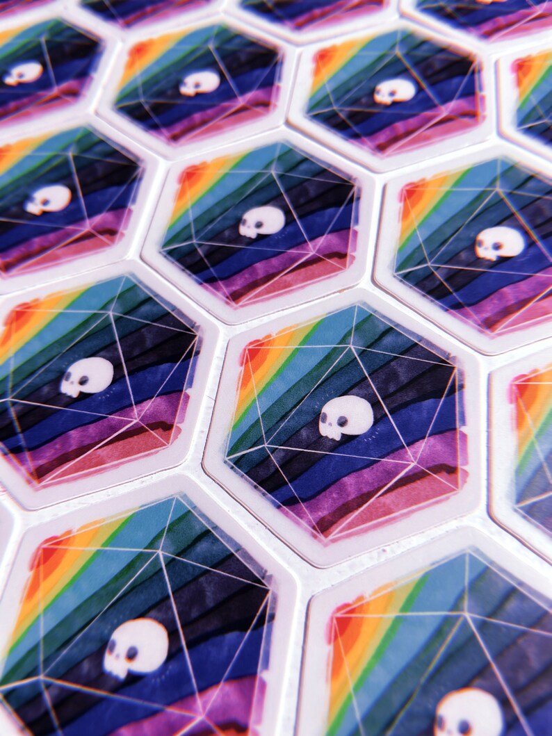 Puede incluir: Primer plano de fichas de juego hexagonales con un dise&ntilde;o de arco&iacute;ris y un gr&aacute;fico de calavera. Las fichas son blancas con un acabado brillante.