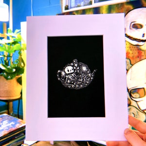 Puede incluir: Una ilustración en blanco y negro de un esqueleto sentado en una tetera con estrellas y otros objetos celestes a su alrededor. La ilustración está enmarcada en un passe-partout blanco.