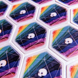 Puede incluir: Un conjunto de fichas hexagonales de juego con un dise&ntilde;o de arco&iacute;ris y un gr&aacute;fico de calavera blanca. Las fichas est&aacute;n hechas de un material transparente y tienen un acabado brillante.