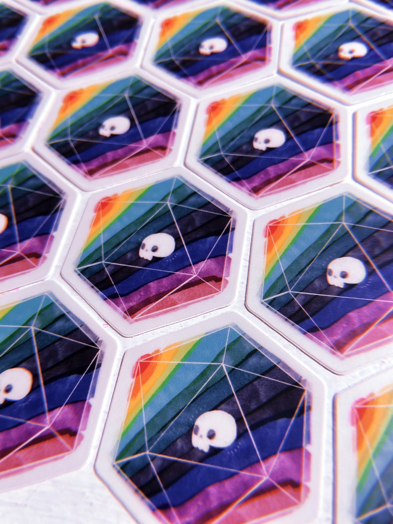 Puede incluir: Un conjunto de fichas hexagonales de juego con un dise&ntilde;o de arco&iacute;ris y un gr&aacute;fico de calavera blanca. Las fichas est&aacute;n hechas de un material transparente y tienen un acabado brillante.