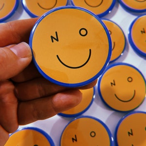 Puede incluir: Una colección de botones amarillos y azules con forma de cara sonriente con la palabra "NO" impresa en ellos. Los botones están dispuestos en una cuadrícula sobre una superficie blanca.