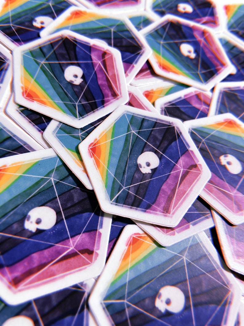 Puede incluir: Una pila de pegatinas hexagonales de colores con un dise&ntilde;o de arco&iacute;ris y una calavera blanca en el centro. Las pegatinas est&aacute;n impresas en papel blanco y tienen un acabado brillante.
