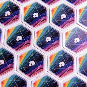 Puede incluir: Primer plano de un conjunto de dados hexagonales con un dise&ntilde;o de arco&iacute;ris y un gr&aacute;fico de calavera. Los dados son blancos con una parte superior e inferior de pl&aacute;stico transparente.