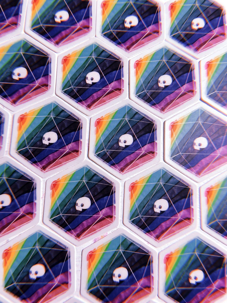 Puede incluir: Primer plano de un conjunto de dados hexagonales con un dise&ntilde;o de arco&iacute;ris y un gr&aacute;fico de calavera. Los dados son blancos con una parte superior e inferior de pl&aacute;stico transparente.