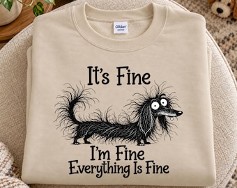May include: A beige crewneck sweatshirt featuring a black graphic of a long-haired, disheveled dachshund with wide eyes and a tongue sticking out. The text "It's Fine, I'm Fine, Everything Is Fine" is printed in black above and below the dog illustration.