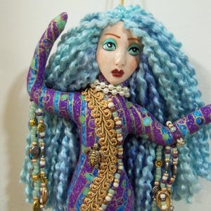 May include: A handmade mermaid doll with blue yarn hair and a painted face. The doll wears a purple and gold patterned dress and is adorned with beaded necklaces and charms. A gold cord is attached for hanging.