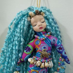 May include: A handmade mermaid doll with blue yarn hair and a painted face. The doll wears a colorful patterned fabric outfit, adorned with seashells, beads, and a turtle charm. A gold string is attached for hanging.