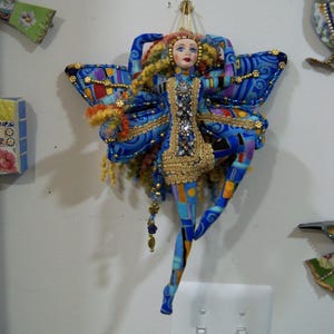 May include: A handcrafted fairy doll with blue butterfly wings, adorned with gold trim and colorful patchwork patterns. The doll has a painted face, curly hair, and beaded accents. The date "18/12/2025" is printed on the wall.