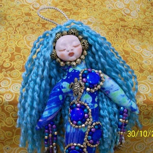 May include: A handmade doll with blue yarn hair and a painted face. The doll is wearing a blue and purple fabric dress adorned with blue and purple jewels and a turtle pendant. The background is a gold patterned fabric.