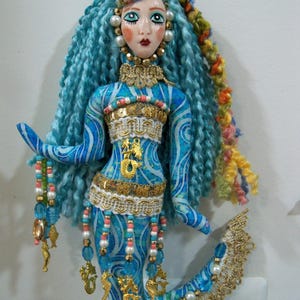 May include: A handcrafted mermaid ornament with blue yarn hair and a painted face. The mermaid's body is adorned with blue and white fabric, gold accents, and beaded embellishments. The ornament is approximately 10 inches tall.