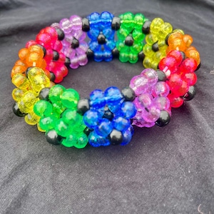 May include: A circular bracelet constructed from translucent, multi-coloured plastic beads arranged in a repeating flower pattern. The design features small black beads at the centre of each flower, with petals in shades of red, orange, yellow, green, blue, and purple.
