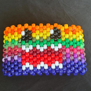 May include: A wide cuff bracelet constructed from rows of colourful plastic pony beads arranged in a rainbow gradient pattern. The design features a central graphic of a face with black eyes and white teeth, created using contrasting bead colours.