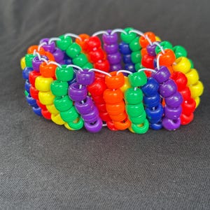May include: A multi-row cuff bracelet constructed from numerous small, cylindrical plastic pony beads in a vibrant rainbow colour palette. The beads are arranged in vertical columns and connected by thin, white elastic cord, creating a wide, flexible band.