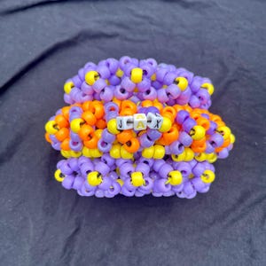 Puede incluir: Una pulsera de brazalete multicapa construida con pequeñas cuentas de plástico en tonos morado, naranja y amarillo. La fila central presenta cuentas cuadradas blancas con letras negras que forman el nombre JAX en mayúsculas.