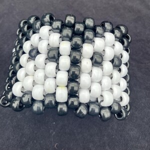 May include: A wide cuff bracelet constructed from rows of glossy black and opaque white plastic pony beads. The beads are arranged in a repeating grid pattern, creating a thick, multi-layered band that maintains a structured, cylindrical shape.