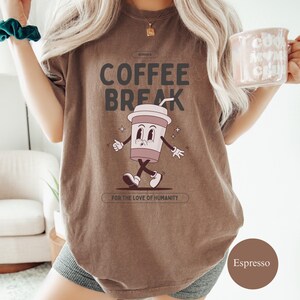 Puede incluir: Camiseta color espresso con las palabras "COFFEE BREAK" y un dibujo de una taza de café de dibujos animados. La taza tiene brazos, piernas y una cara sonriente. El texto "FOR THE LOVE OF HUMANITY" está debajo.