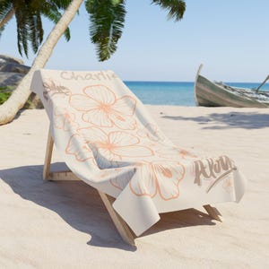 May include: A beach chair with a cream-colored towel featuring a floral design in peach tones. The towel has the name "Charlie" and the word "Aloha" printed on it. The chair is on a sandy beach with a boat in the background.