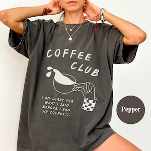 May include: Charcoal grey t-shirt featuring the words "COFFEE CLUB" in white, a coffee cup graphic, and the text "I AM SORRY FOR WHAT I SAID BEFORE I HAD MY COFFEE:)". The shirt has short sleeves and a relaxed fit.