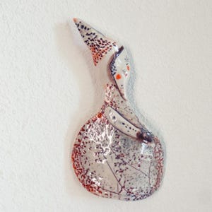 May include: A ceramic wall sculpture of a bird. The artwork is mainly white with speckled patterns in black and red. The bird features a long, pointed beak and small orange accents, mounted on a white wall.