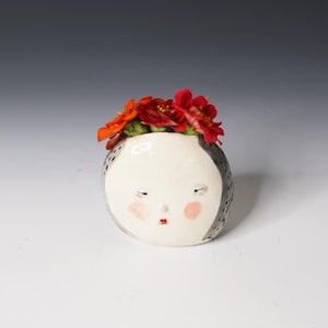 May include: A round white ceramic vase shaped like a face with closed eyes, a small nose, and pink cheeks. The sides are dark gray with a dotted pattern. The top is filled with a cluster of vibrant red and orange artificial flowers.