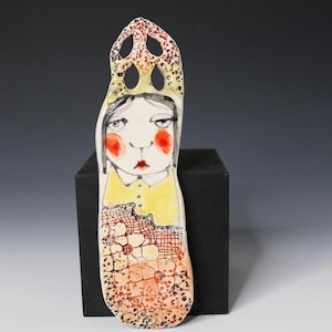 May include: A tall, flat ceramic wall hanging featuring a stylized illustration of a person with dark hair, bright red cheeks, and a yellow collared shirt. The top has a cutout design, and the lower section displays a textured orange grid pattern.