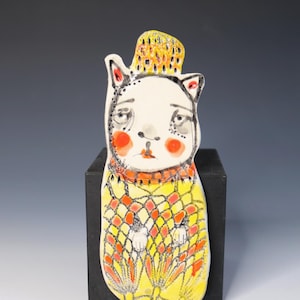 May include: A flat ceramic wall hanging shaped like a cat with a white face, red cheeks, and a yellow hat. The body features a yellow and orange grid pattern with small white paws visible. The cat has black outlined features and a yellow base.