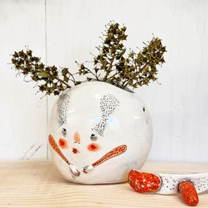 May include: A round white ceramic vase featuring a hand-painted face with red cheeks, black dotted ears, and orange arms. The vase contains dried brown plant stems. A separate white ceramic piece with orange textured accents sits in front.
