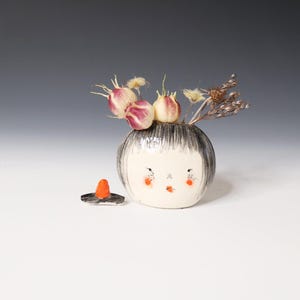 May include: A round white ceramic vase shaped like a head with black painted hair, orange cheeks, and a small mouth. The vase contains dried flowers and is accompanied by a small, separate black and orange conical ceramic piece.