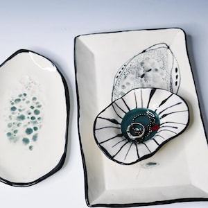 May include: Three ceramic dishes with off-white glaze and black painted edges. One oval dish features teal speckled patterns, a rectangular tray displays a black-lined abstract shape, and a small scalloped bowl contains a silver ring.