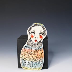 May include: A flat ceramic figurine shaped like a person with a white face, red cheeks, and dark eyes. The figure wears a grey ruffled collar and a body decorated with a textured, dotted pattern in shades of orange, yellow, and blue.