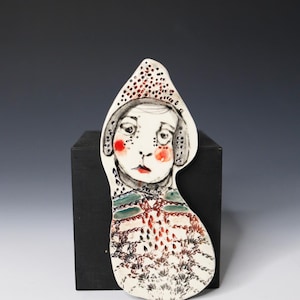 May include: A flat, irregularly shaped ceramic wall hanging featuring a hand-painted illustration of a figure wearing a pointed hood. The design includes red cheeks, dark outlined facial features, and a textured, patterned body in shades of green, red, and black.