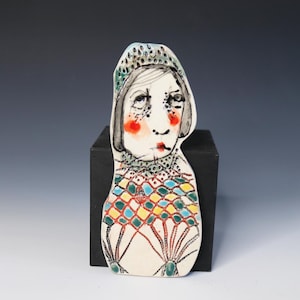 May include: A flat ceramic figurine featuring a stylized portrait of a person wearing a patterned head covering and a high-collared garment. The piece is decorated with a colorful grid pattern in blue, yellow, and red, with black line details.