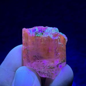 Könnte beinhalten: Ein durchscheinendes, orange-rosa Mineralexemplar mit rauer, strukturierter Oberfläche. Die Oberseite des Minerals weist grüne und blaue Farbtöne auf. Der Hintergrund ist dunkelblau und erzeugt einen Kontrast.