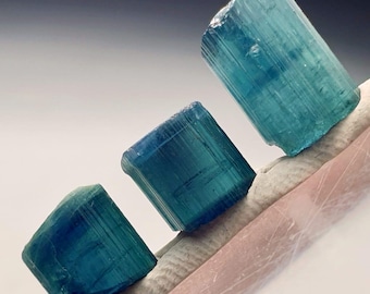 Puede incluir: Se muestran tres cristales de turmalina azul-verdosa. Los cristales rectangulares tienen estrías visibles, descansando sobre una superficie clara y texturizada. Los cristales varían ligeramente en tamaño y tono, mostrando la belleza natural de la gema.