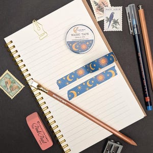 May include: A stationery flat lay featuring a spiral-bound notebook, washi tape, pencils, stamps, and an eraser. The washi tape has a blue background with a sun, moon, and star pattern. The notebook is open to a lined page.